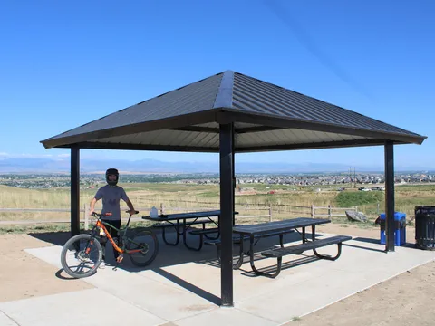 The shelter at the Erie Singletrack trailhead on a clear day, a rider with a mountain bike beside it, and the town and the Front Range spread out below